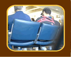 picture of a man sitting in an airport seat. He is cuffed and escorted by law enforcement officials